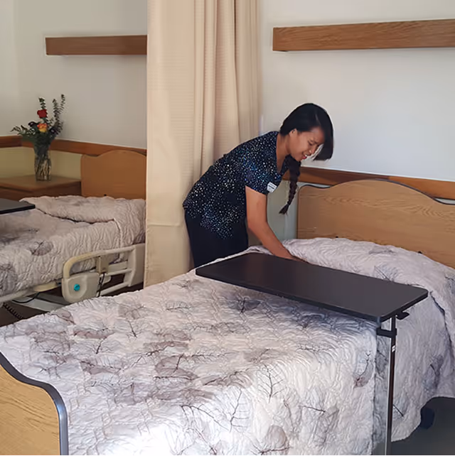 A caregiver making a bed in a healthcare facility room with two hospital-style beds, each with a light-colored quilted blanket. There is a small table over one bed and a vase with flowers on a nightstand between the beds.