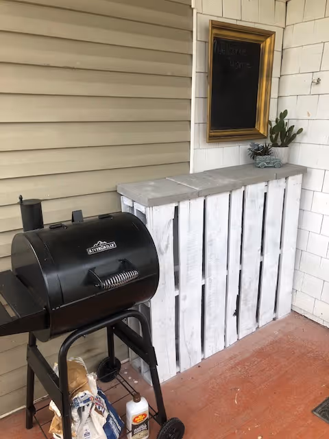 A covered outdoor porch area with a black RiverGrille smoker grill on wheels, a white wooden pallet-style counter with a concrete top, a small potted cactus, and a chalkboard with a gold frame hanging on the wall that says 'Welcome Home'.