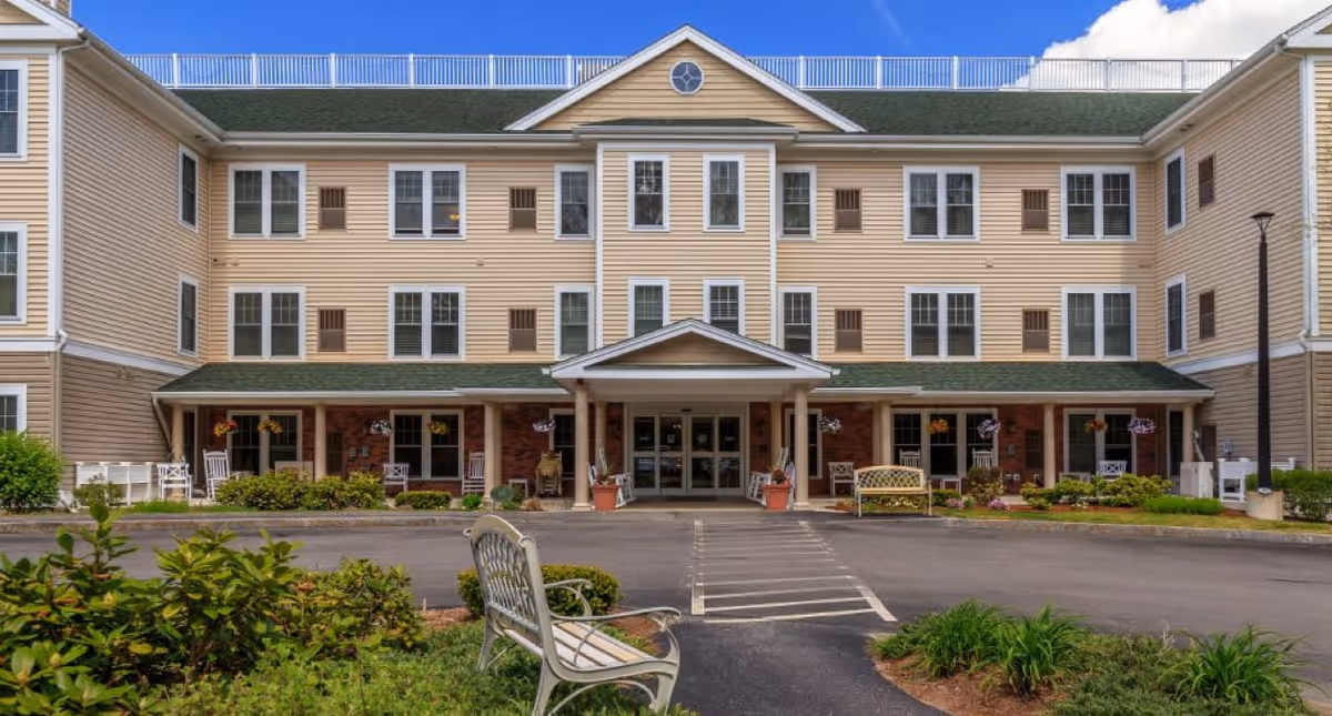 Front exterior view of a three-story senior living facility building with beige siding and green roof. The entrance has a covered porch with white columns, benches, and potted plants. There are multiple windows and some rocking chairs on the porch. The foreground shows a paved driveway and landscaped greenery with benches.