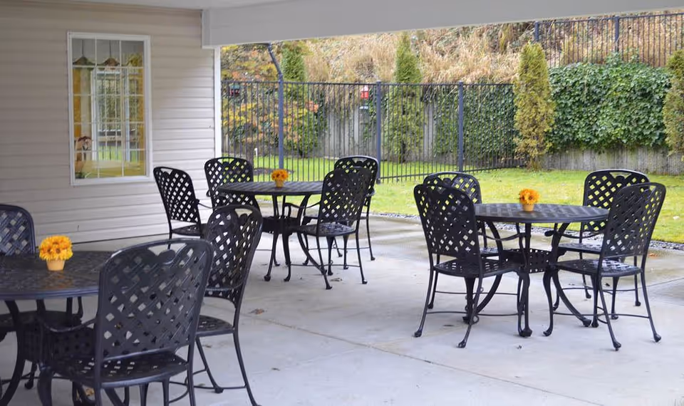 Outdoor covered patio area with several black metal tables and chairs arranged on a concrete floor. Each table has a small yellow flower centerpiece. In the background, there is a grassy area with a black metal fence and some greenery along the fence line.