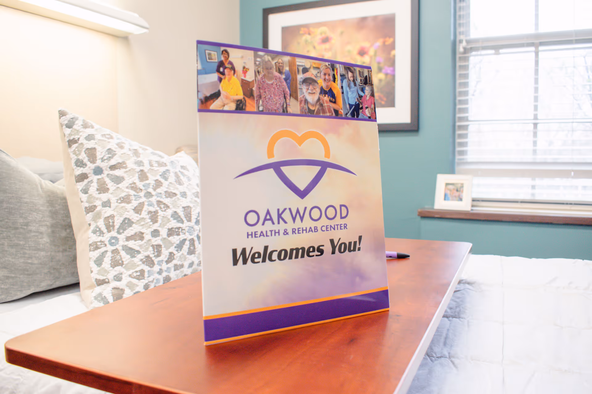 A welcome sign for Oakwood Health & Rehab Center stands on a wooden overbed table in a bright bedroom with pillows and a window.