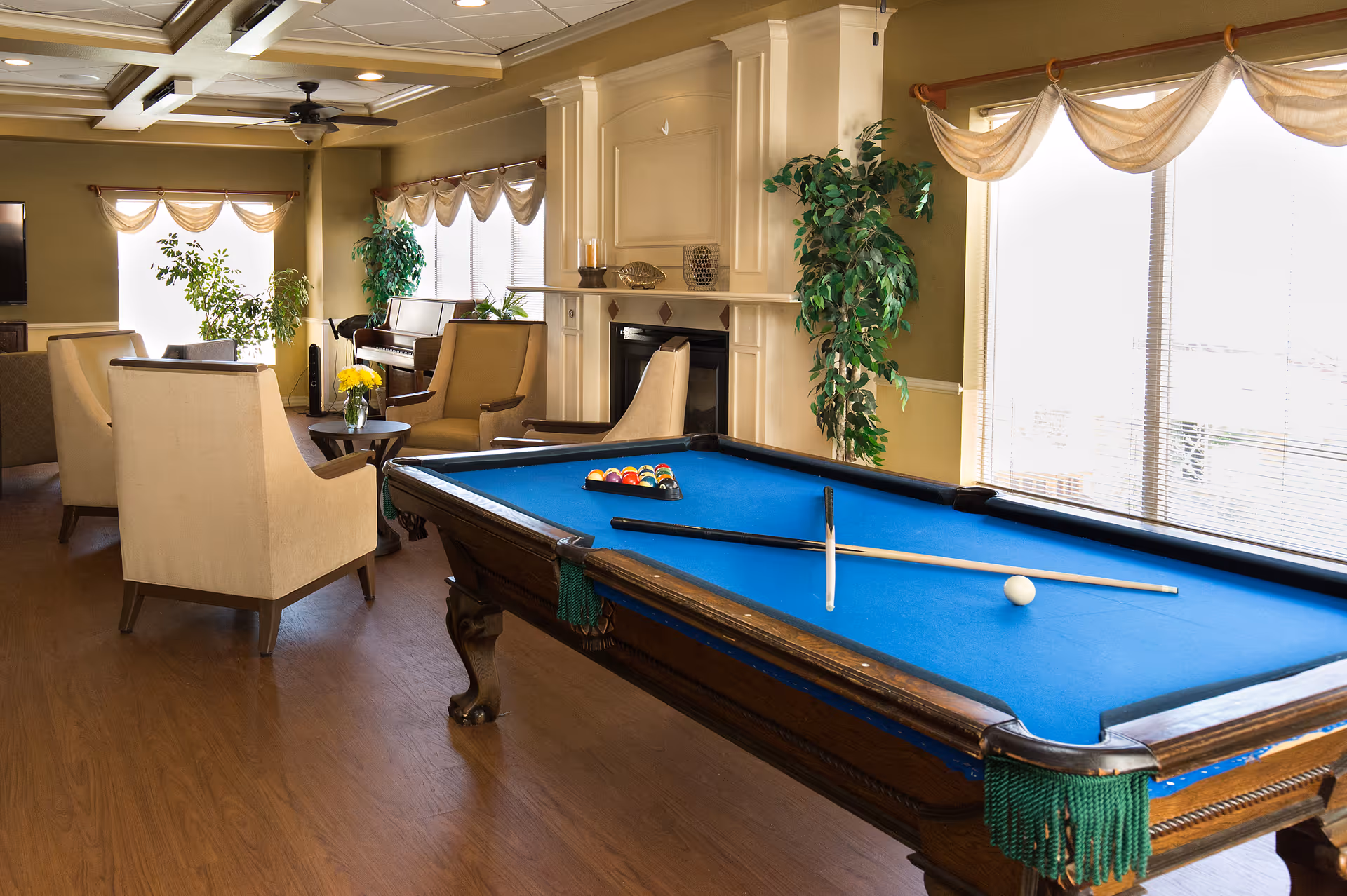 A well-lit living room area with a blue felt pool table in the foreground, surrounded by beige armchairs and small tables. The room features large windows with sheer curtains, a fireplace, potted plants, and a piano in the background.