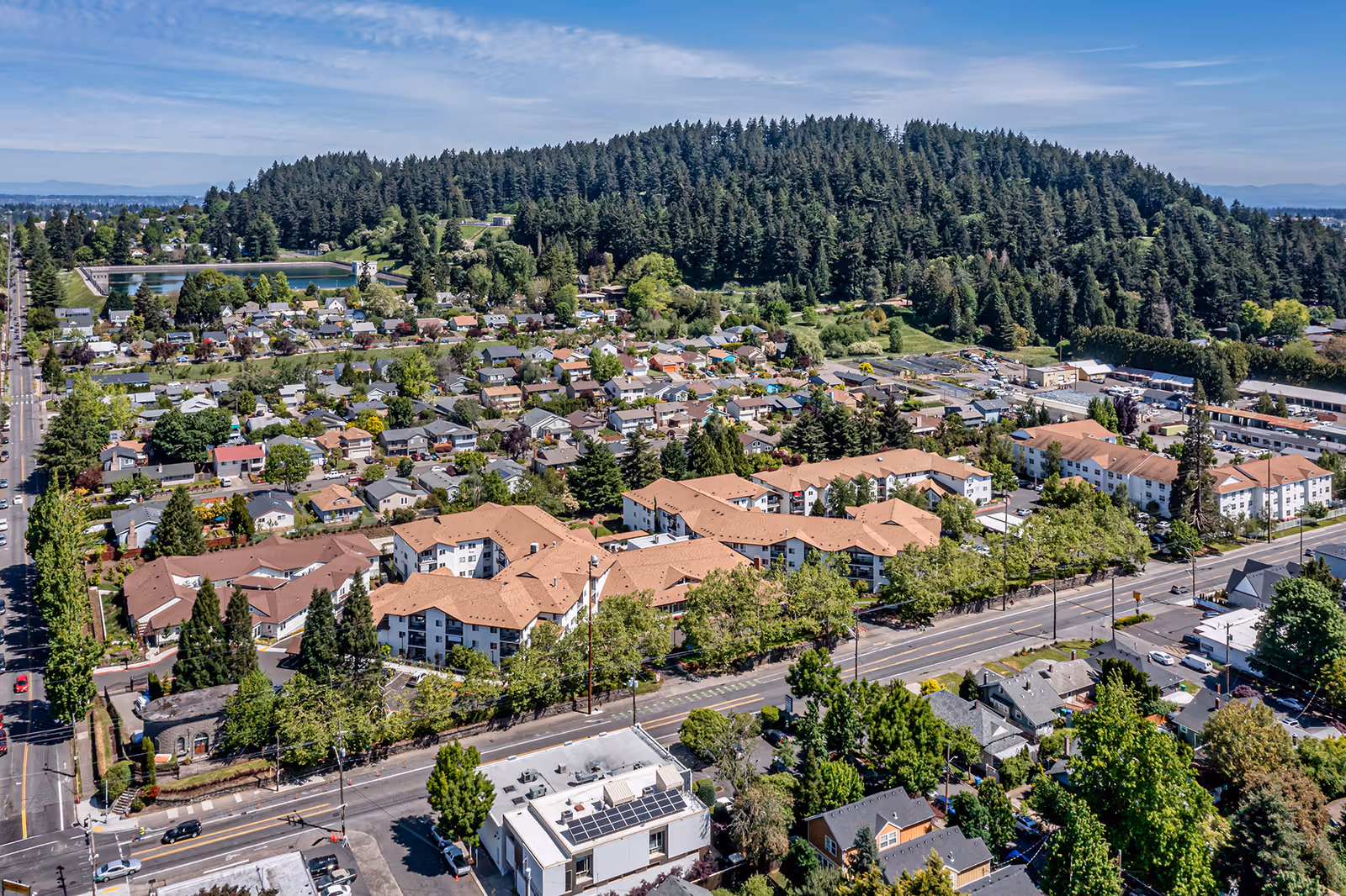 Aerial view of a residential neighborhood with a large senior living facility named Courtyard At Mount Tabor featuring multiple connected buildings with brown roofs, surrounded by trees and streets. In the background, there is a forested hill and a water reservoir under a partly cloudy blue sky.