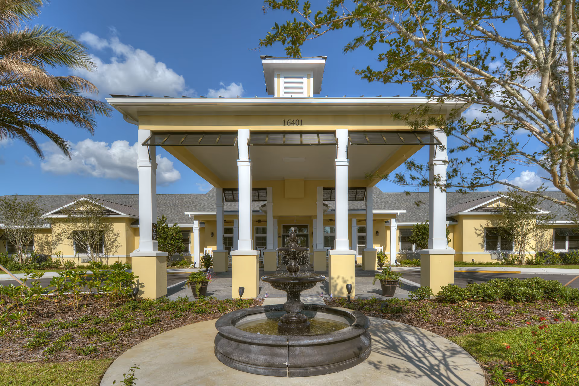 Front exterior view of Benton House of Clermont featuring a covered entrance with white columns, a central water fountain, landscaped garden beds, and a clear blue sky with some clouds.