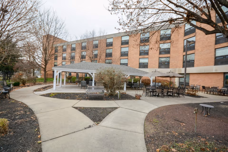 Outdoor courtyard area of a senior living facility with a covered pavilion, multiple tables with chairs and umbrellas, benches, and a brick multi-story building in the background. Leafless trees and paved walkways surround the seating areas.