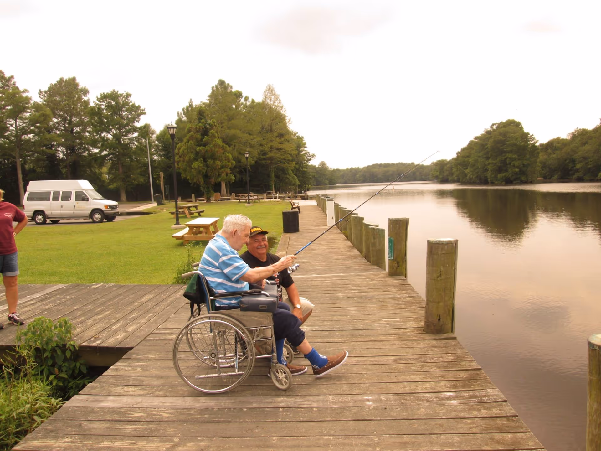 An elderly man in a wheelchair fishing off a wooden dock by a calm river, accompanied by another man kneeling beside him. In the background, there is a grassy area with picnic tables, trees, and a white van parked on a paved area.
