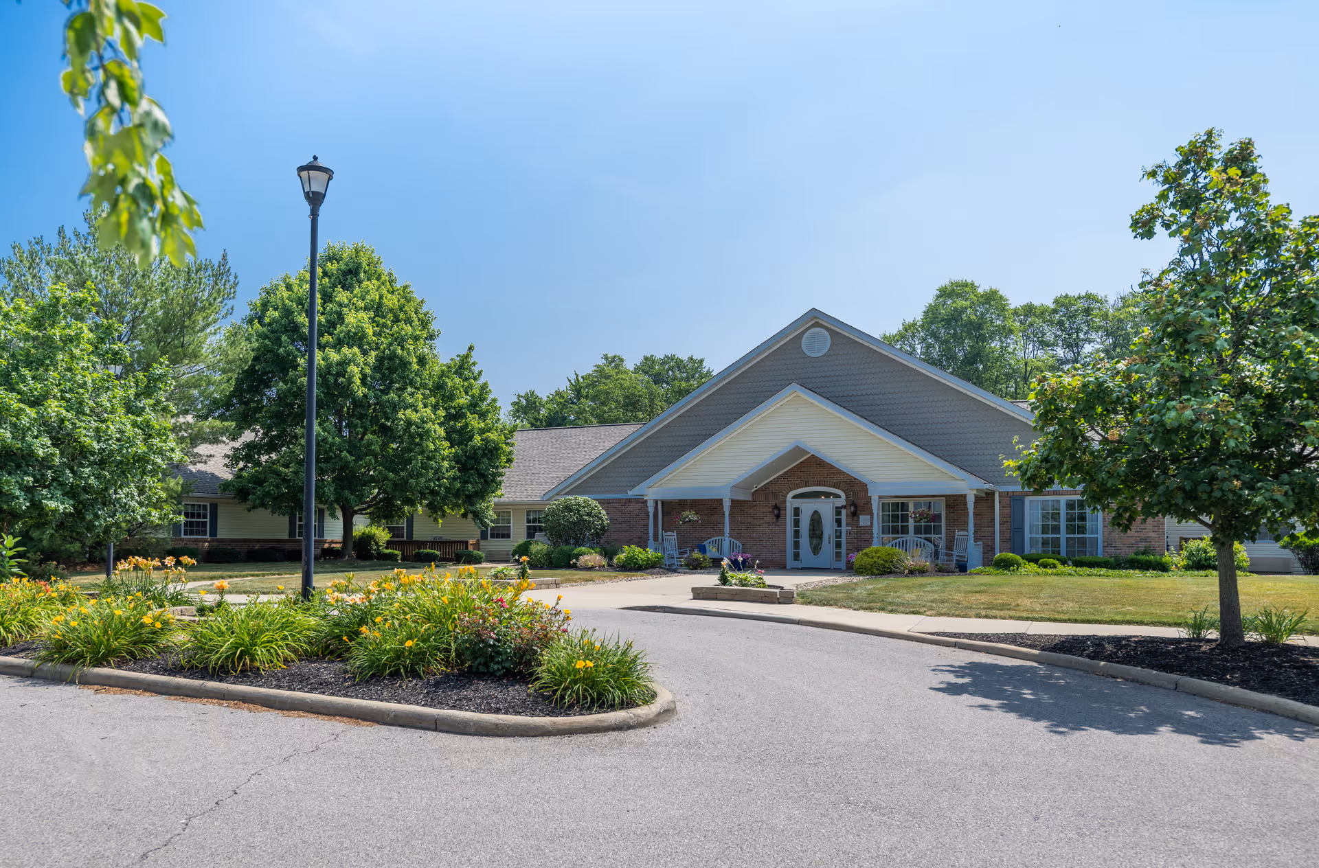 Front entrance of a single-story brick senior living building with a circular driveway, lamp post, and landscaped flower beds.