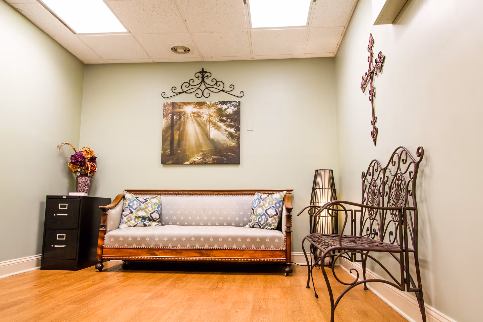 A cozy sitting area with a wooden framed sofa with patterned cushions, a black metal decorative bench, a tall floor lamp, a small black filing cabinet with a vase of flowers on top, and a wall decoration featuring a cross and a framed picture of sunlight streaming through trees.