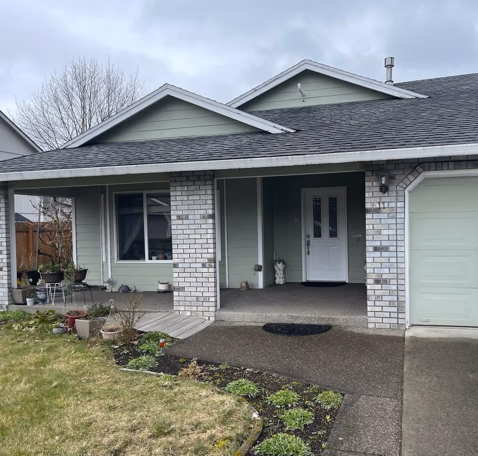 Front exterior of a single-story house showing a covered porch with brick pillars, a white entry door, attached garage, and a small front yard.