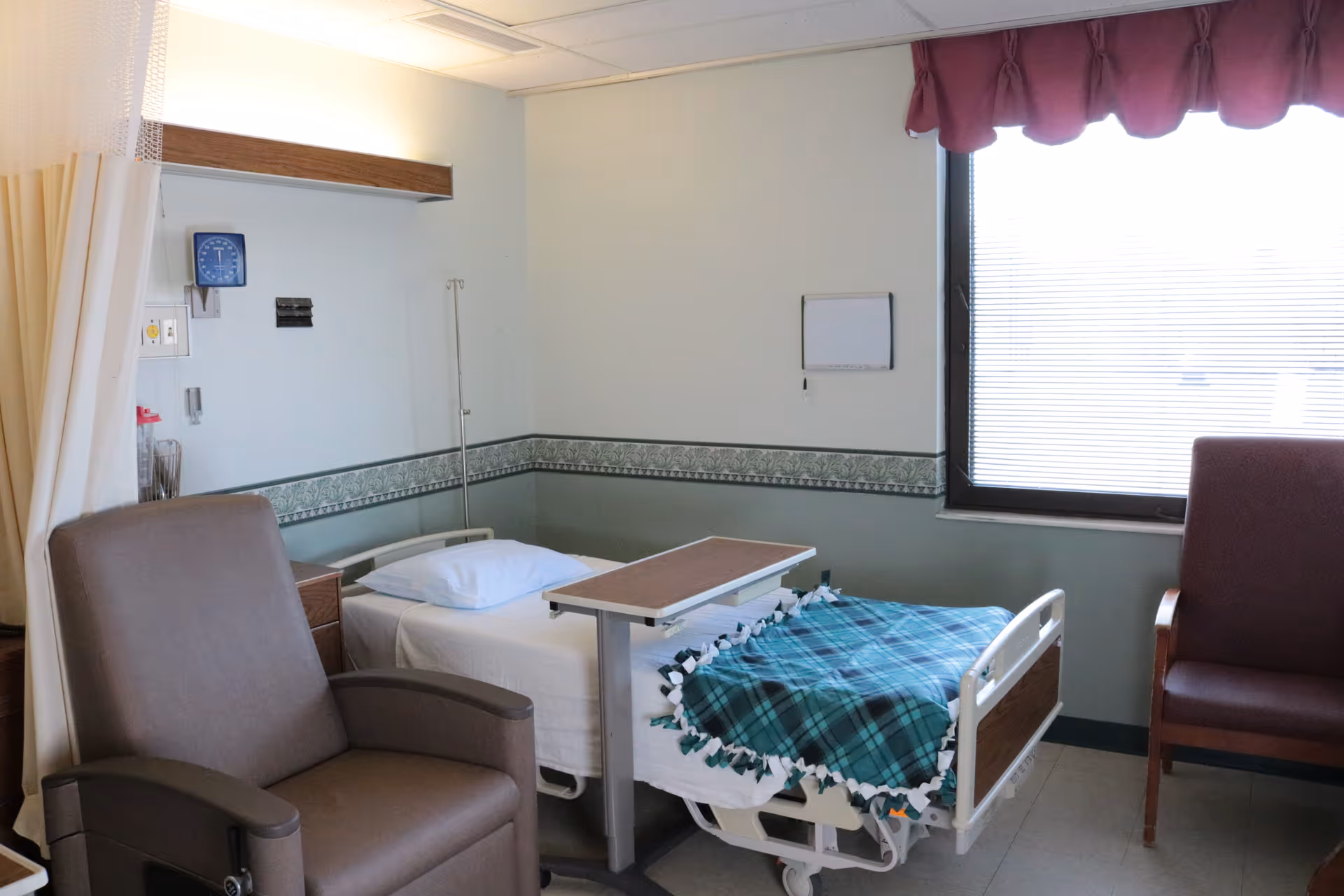 A bright patient room with a hospital bed covered by a plaid blanket, an overbed table, reclining chairs, an IV pole, and a window with blinds.