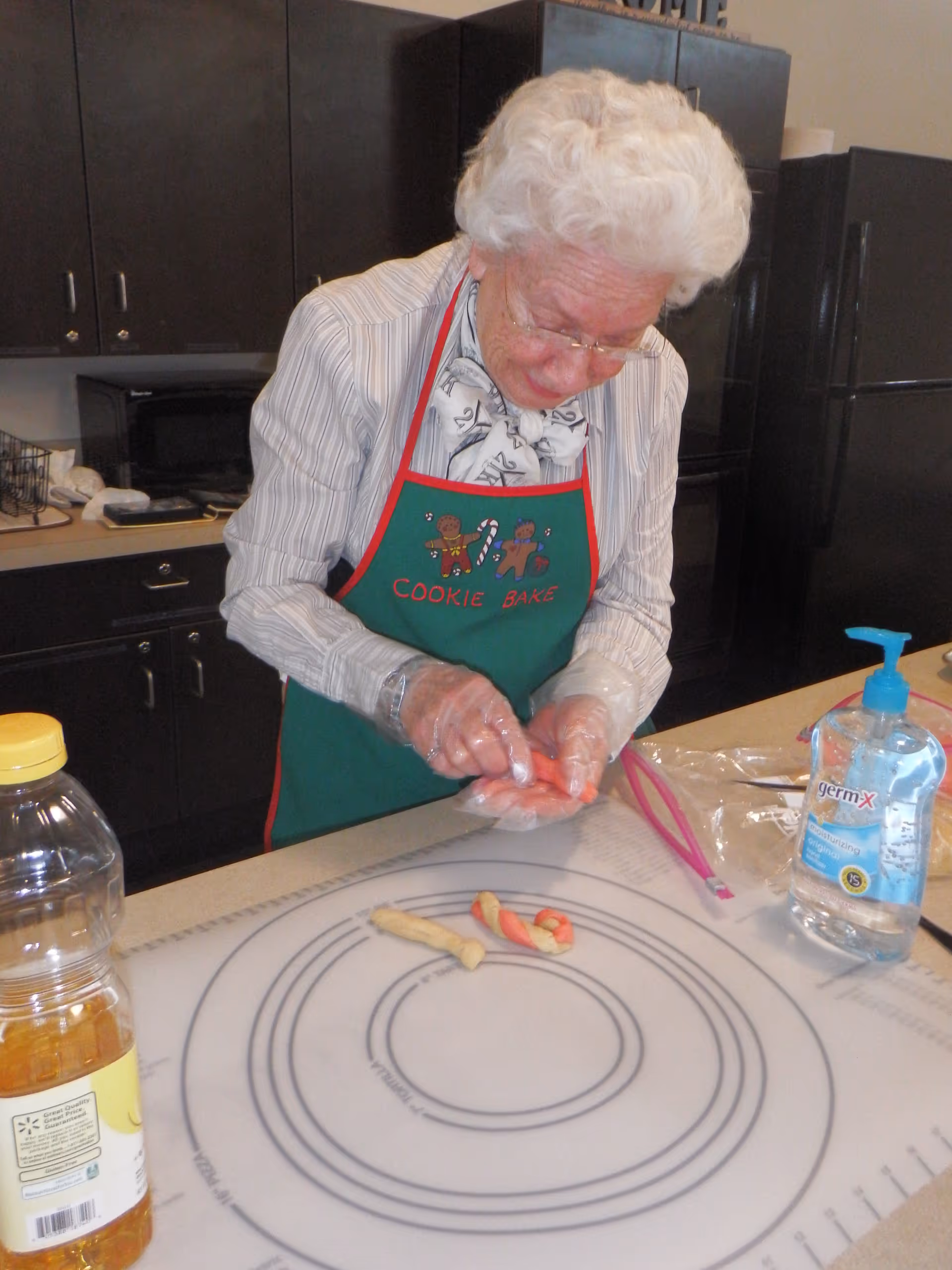 An elderly woman wearing a green apron with gingerbread men and the words 'COOKIE BAKE' is shaping dough on a kitchen counter. She is wearing gloves and standing in a kitchen with dark cabinets and a black refrigerator. There is a bottle of cooking oil and a bottle of Germ-X hand sanitizer on the counter.