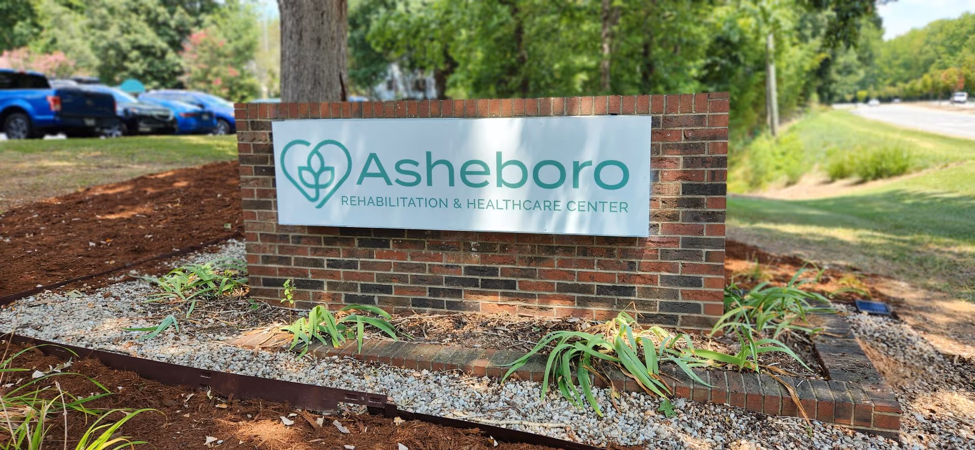 A brick sign for Asheboro Rehabilitation & Healthcare Center situated outdoors with some plants and gravel around its base. There are trees, grass, and parked cars visible in the background along a road.