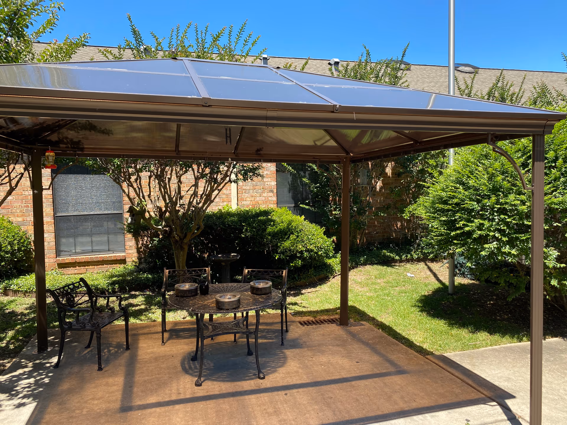 Outdoor covered patio with a metal gazebo, table and chairs in a garden courtyard next to a brick building.