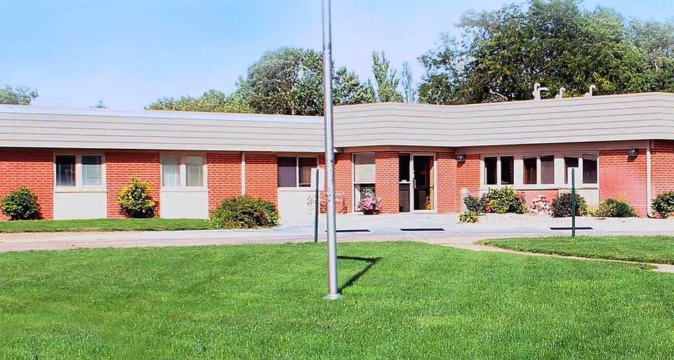 Single-story brick building with windows and entrance, a flagpole in front, and a green lawn.