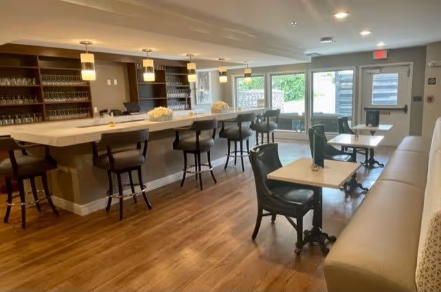 A bright and modern dining area in a senior living facility featuring a long bar counter with high chairs, several small tables with chairs, wooden flooring, and large windows allowing natural light to enter.