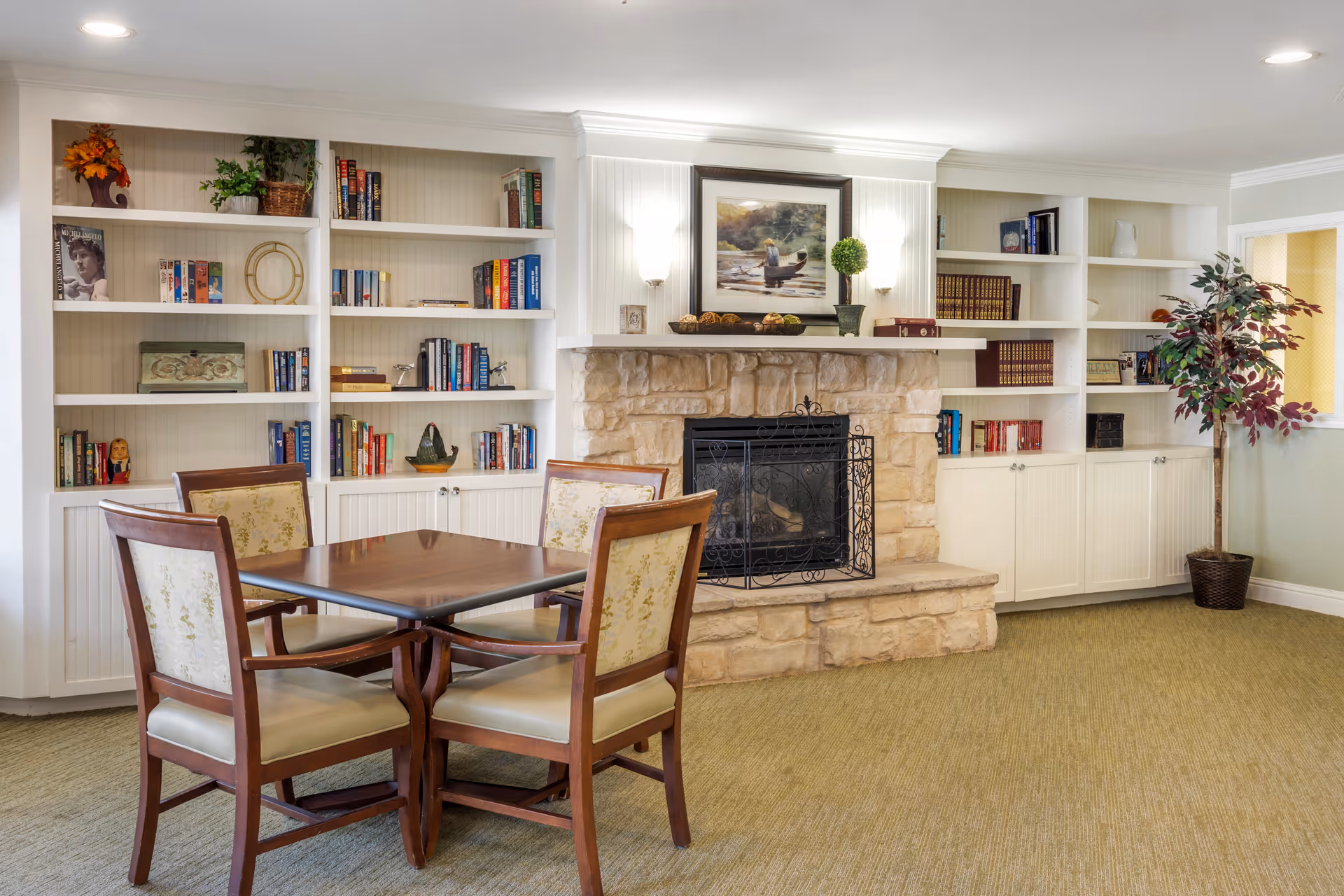 A cozy room with a stone fireplace in the center, flanked by white built-in bookshelves filled with books and decorative items. In front of the fireplace is a wooden table with four upholstered chairs. A framed painting hangs above the fireplace, and a potted plant stands to the right side of the room. The room has a carpeted floor and soft lighting.