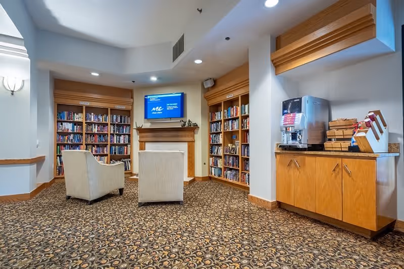 A cozy library or reading area in a senior living facility with two beige armchairs facing a wall-mounted TV above a wooden mantel. The room has built-in wooden bookshelves filled with books on either side of the mantel. To the right, there is a wooden cabinet with a coffee machine and supplies on top. The floor is covered with a patterned carpet, and the walls are painted light with wooden trim.