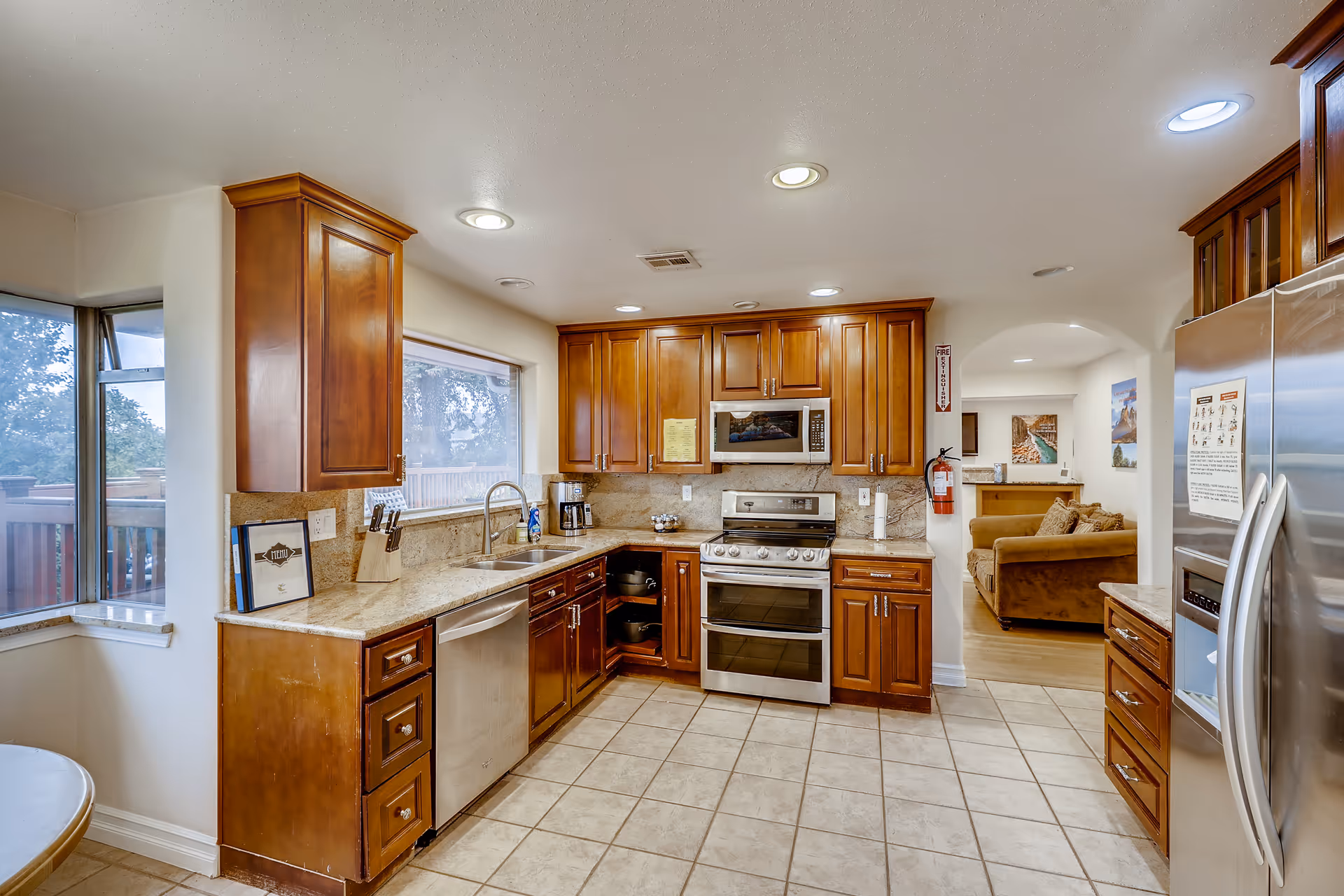 Bright kitchen with wooden cabinets, granite countertops, stainless steel appliances, and a tiled floor opening into a living area.