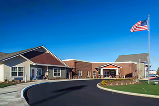Exterior of Marquis Mill Park senior living facility with entrance, curved driveway, flagpole, and lawn.