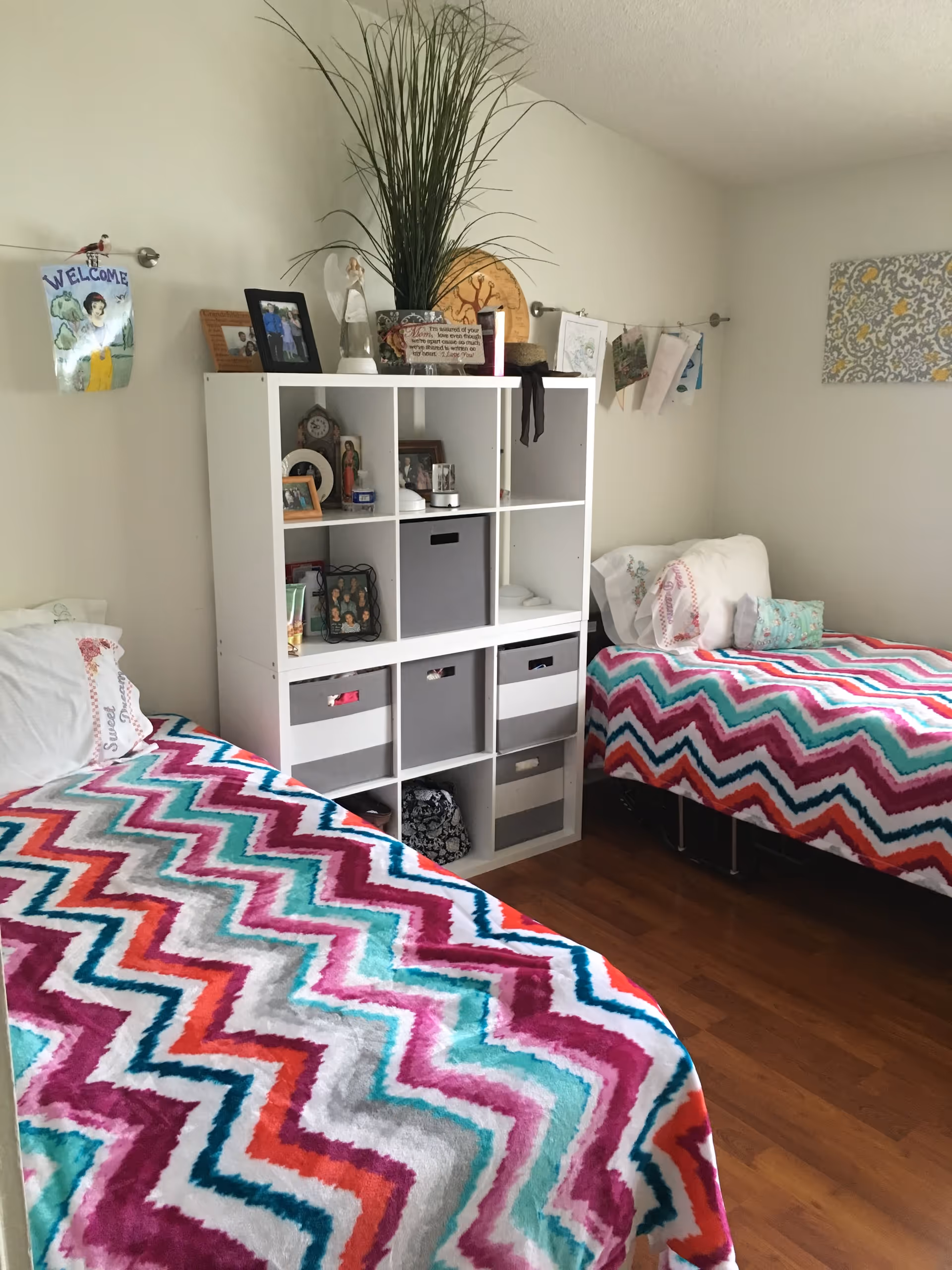 A cozy bedroom with two twin beds covered in colorful zigzag patterned blankets in shades of pink, purple, teal, and white. Between the beds is a white cubical shelving unit holding various decorative items, framed photos, and storage bins. The walls are light-colored with some artwork and a welcome sign hanging above the shelving unit. The floor is wooden.