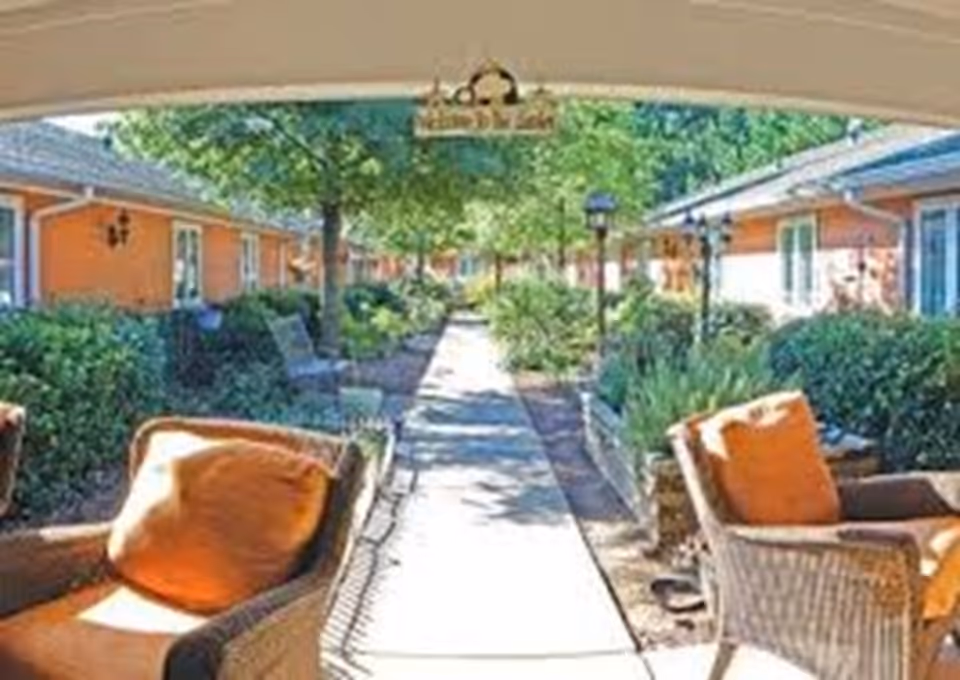 Outdoor seating area with wicker chairs and orange cushions under a covered patio, overlooking a walkway lined with bushes, trees, and orange buildings on both sides.