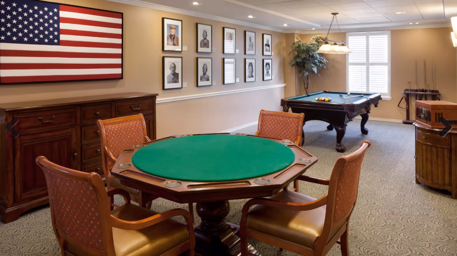 A cozy game room in a senior living facility featuring a green felt octagonal card table with four wooden chairs around it. In the background, there is a pool table with billiard balls and cues, a large American flag on the wall, framed black and white portraits, a wooden cabinet, a potted plant, and a window with white shutters letting in natural light.