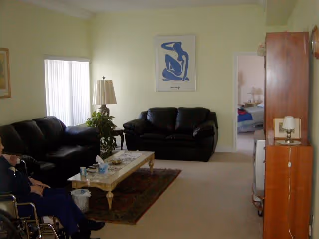 A living room with two black leather sofas, a wooden coffee table with items on it, a floor lamp, a plant, and a framed artwork on the wall. A person in a wheelchair is partially visible on the left side. There is an open doorway leading to a bedroom in the background.
