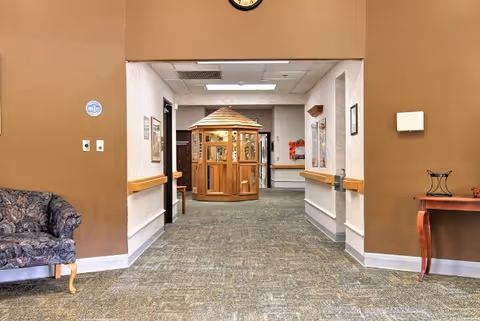 Interior hallway of a healthcare facility with beige walls and carpeted floor. A wooden gazebo-like structure is visible at the end of the hallway. On the left side, there is a patterned upholstered chair, and on the right side, a wooden table with a decorative item. A clock is mounted on the wall above the hallway entrance.