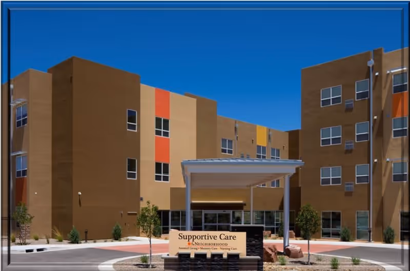 Exterior view of a multi-story building with a covered entrance labeled Supportive Care at The Neighborhood in Rio Rancho Life Plan Community, featuring a clear blue sky and some small trees and landscaping in front.