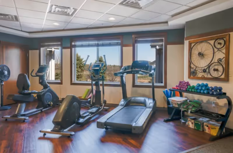 A fitness room with exercise equipment including a recumbent bike, an elliptical machine, and a treadmill. There are large windows showing an outdoor view with trees. On the right side, there is a rack with colorful dumbbells, yoga mats, and other fitness accessories. A decorative wall art piece featuring bicycle wheels is mounted on the wall.