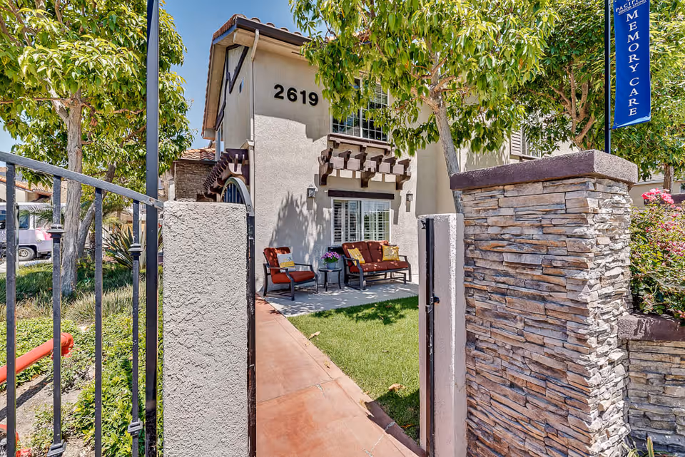 View through an open gate of a senior living facility entrance with a stone pillar on the right and trees on both sides. A two-story building with the number 2619 is visible, featuring a small patio area with outdoor seating including a bench and two chairs with cushions. A blue vertical banner on the right reads 'Pacifica Senior Living Memory Care'.