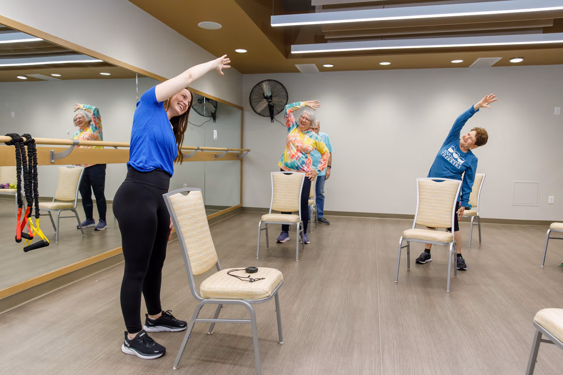 A fitness instructor leads a group of elderly women in a seated stretching exercise in a bright room with wooden floors, beige walls, and a large mirror on one side. The women are standing beside chairs and stretching their arms overhead to the side.