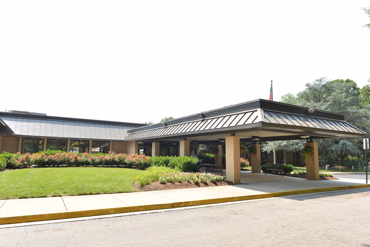 Exterior view of Laurels of University Park facility showing a covered entrance with brick pillars, a manicured lawn, flower beds, and trees in the background under a clear sky.