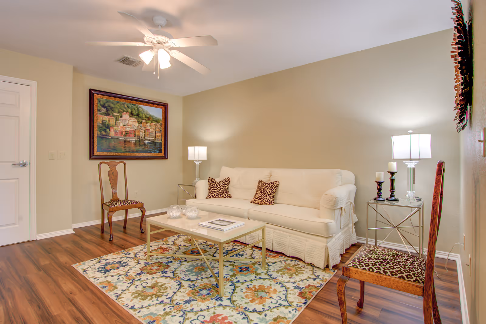 A cozy living room with a white sofa adorned with two patterned pillows, a glass coffee table on a colorful floral rug, two wooden chairs with leopard print cushions, two side tables with lamps and candles, a ceiling fan with lights, and a framed painting of a waterfront village on the wall.