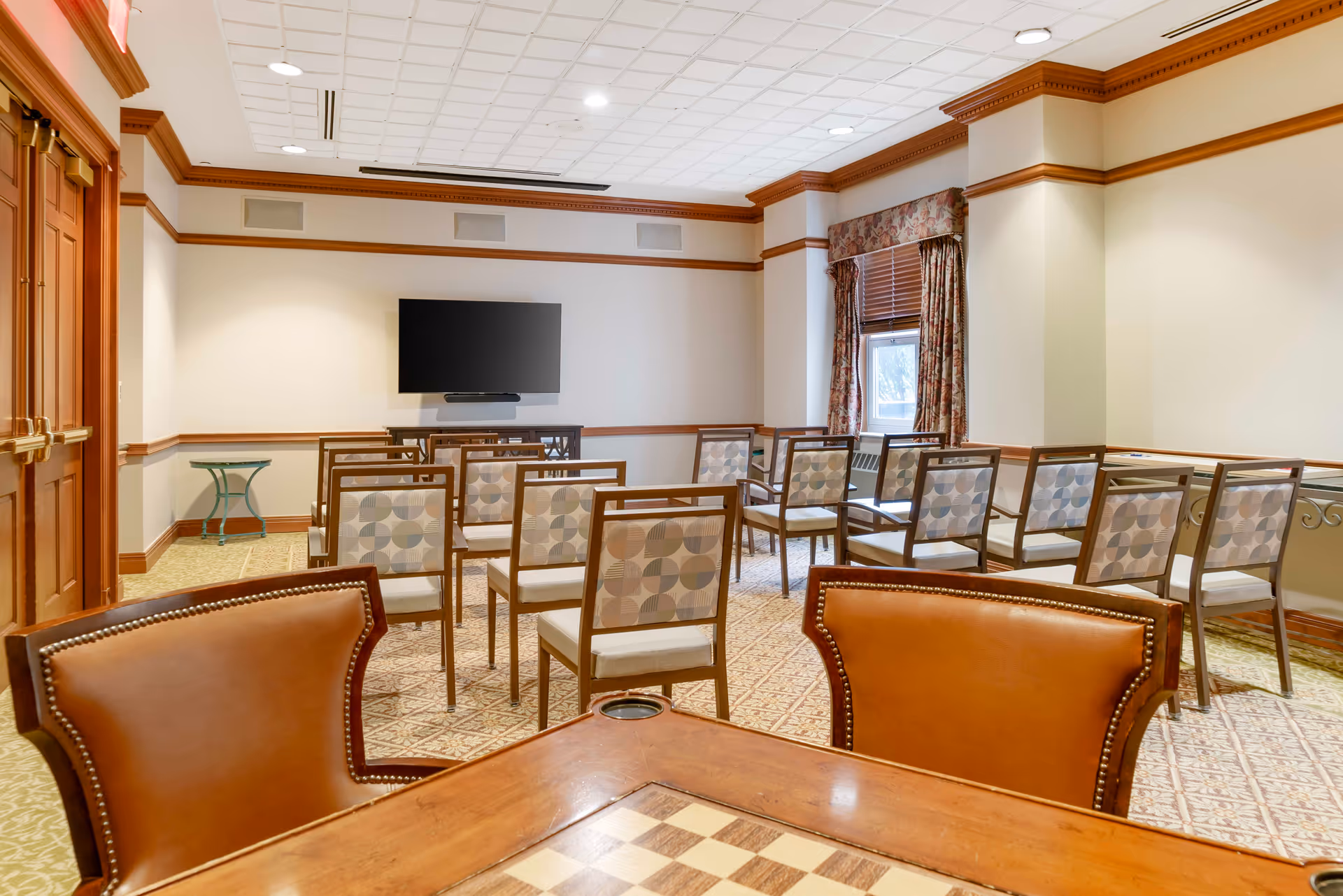 A meeting or activity room with rows of chairs facing a wall-mounted flat screen TV. The room has beige walls with wooden trim, a window with patterned curtains, and a table with a chessboard in the foreground.