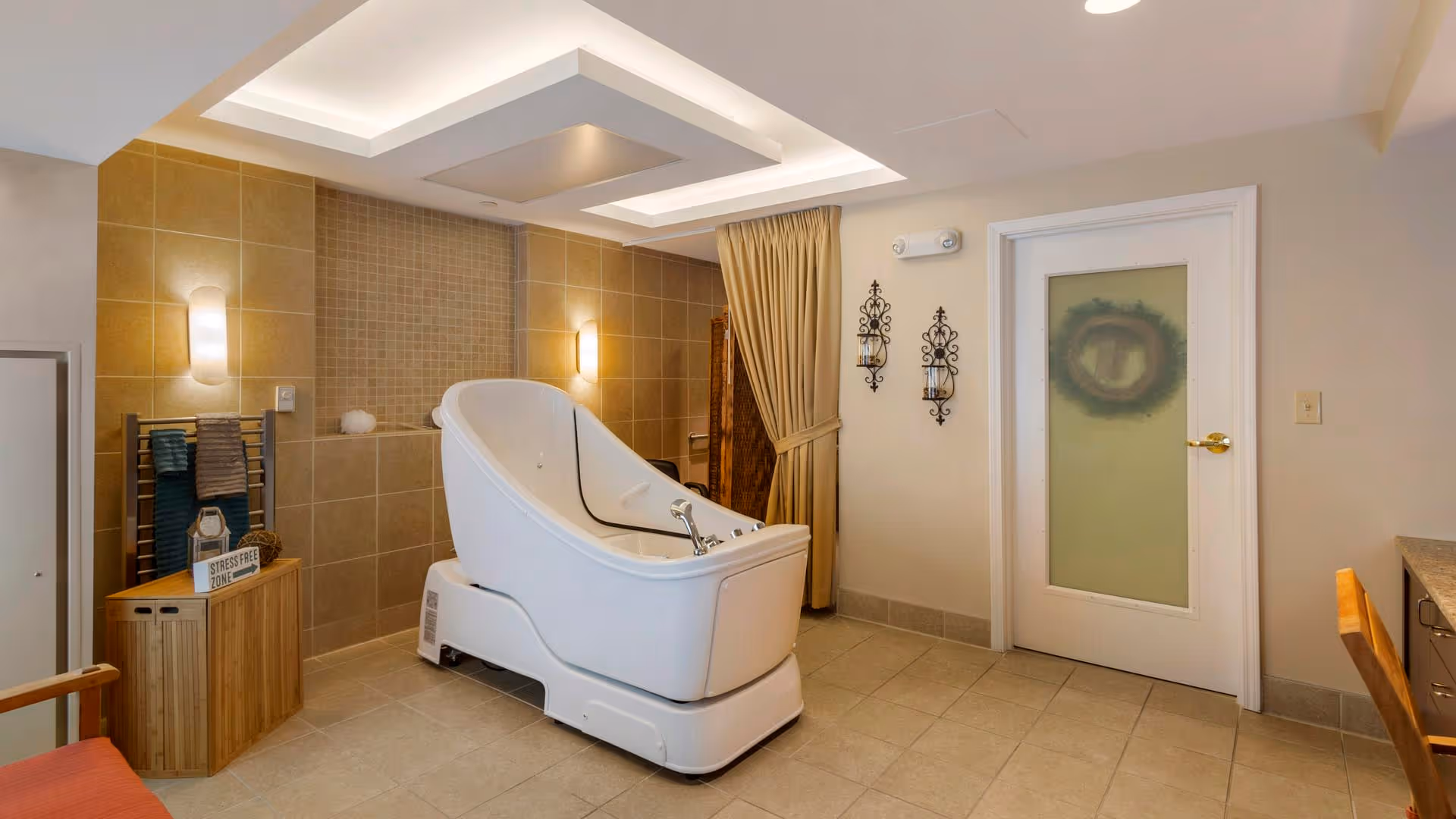 A modern bathing room featuring a large white therapeutic bath tub in the center, beige tiled walls and floor, a wooden towel rack with folded towels, a small wooden cabinet with a 'Stress Free Zone' sign, two wall-mounted candle holders, a beige curtain, and a door with frosted glass.
