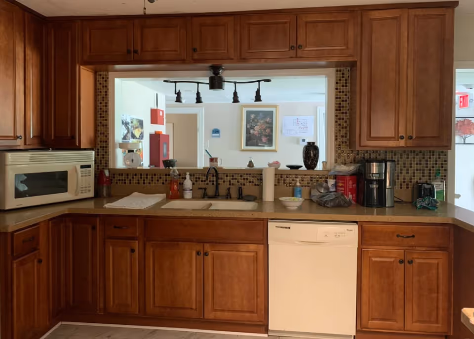 Kitchen area with wooden cabinets, a microwave on the left countertop, a sink with a faucet in the center, a dishwasher below the sink, and a coffee maker on the right countertop. There is a pass-through window above the sink showing a hallway with a framed floral picture and a floor plan on the wall.