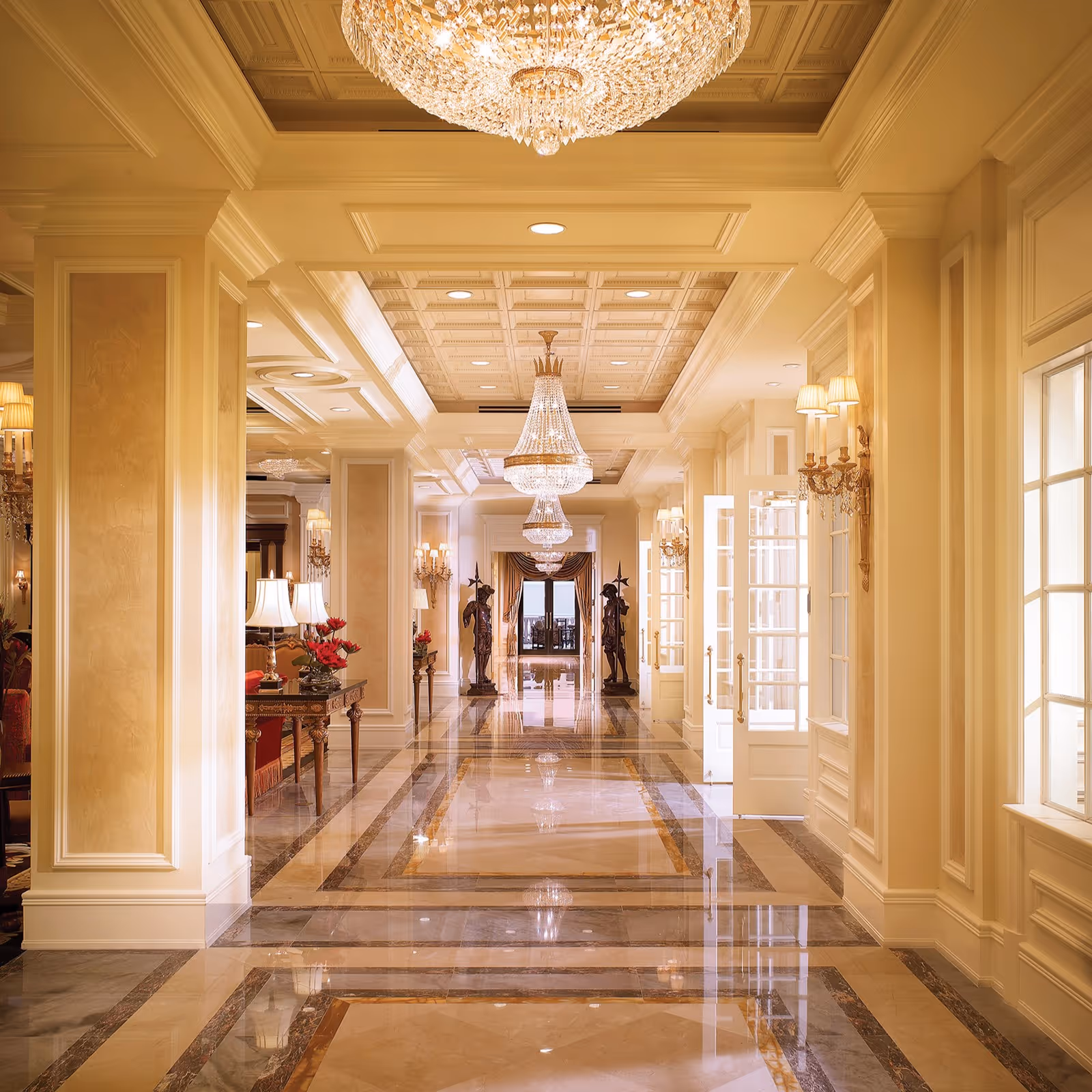 A luxurious hallway with polished marble floors featuring intricate patterns, cream-colored walls with decorative molding, ornate wall sconces, and large crystal chandeliers hanging from a coffered ceiling. At the end of the hallway, two statues flank an open doorway with a view of an outdoor area.