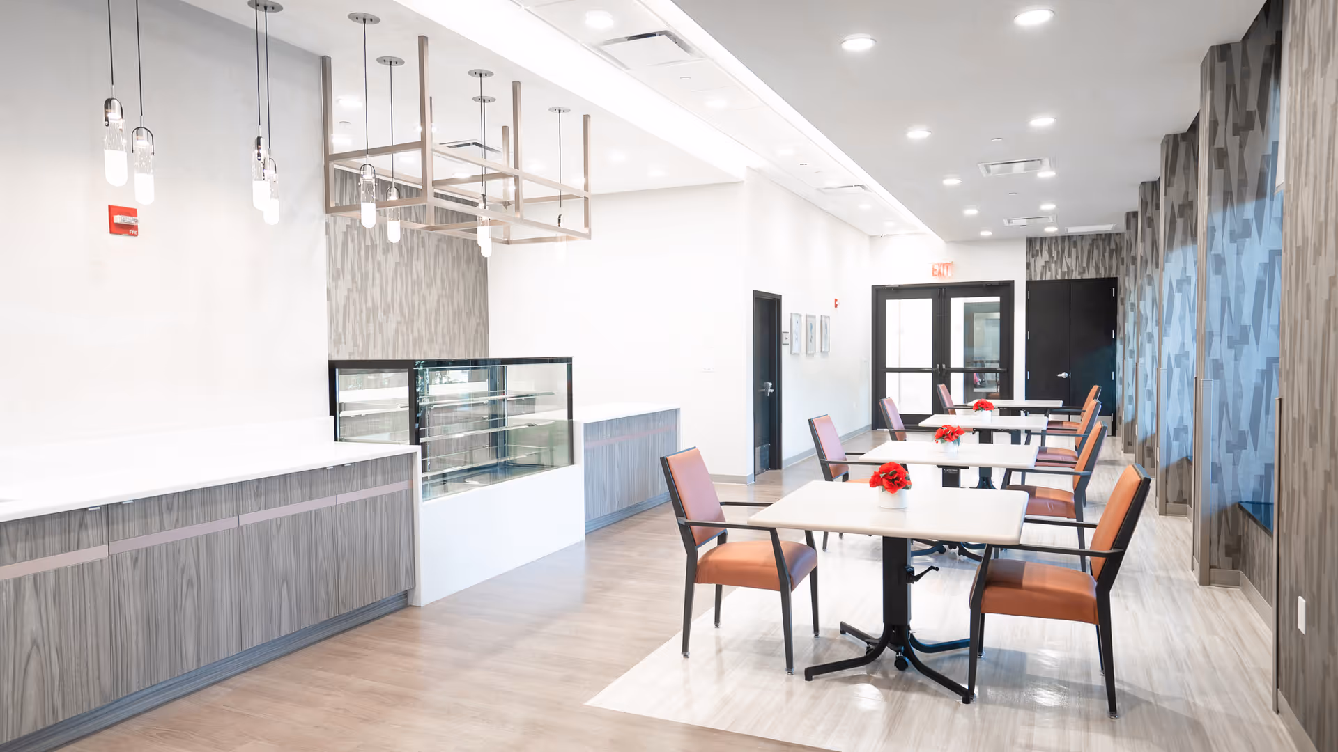 A bright and modern dining area with several tables and chairs arranged neatly. Each table has a small red flower centerpiece. The room features light wood flooring, pendant lights hanging from the ceiling, and a counter with a glass display case. The walls are decorated with patterned wallpaper and there are large windows on one side.