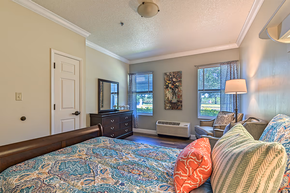Bright, furnished bedroom with patterned bedding, dresser, armchair and two windows.