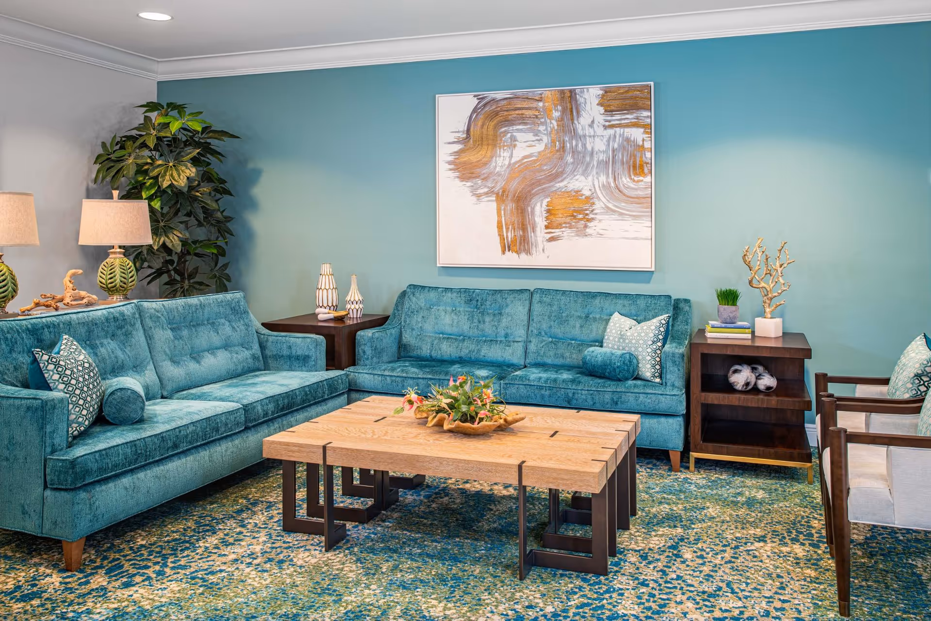 A cozy living room with teal blue sofas, a wooden coffee table with a decorative centerpiece, a patterned rug in shades of blue and green, a side table with lamps, a large potted plant, and abstract artwork on a teal wall.