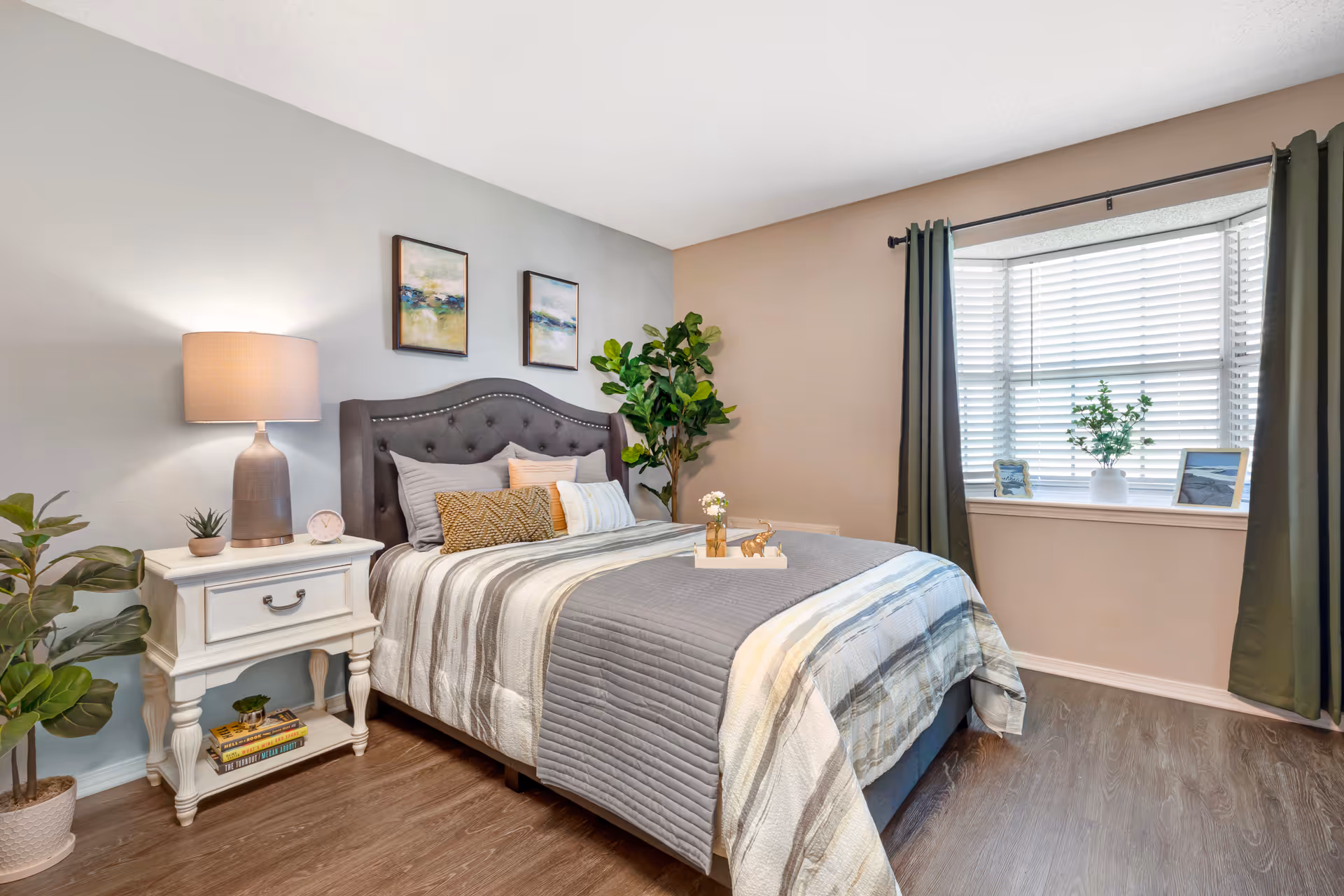 A cozy bedroom with a neatly made bed featuring a gray and white striped comforter and multiple pillows. There is a white nightstand with a lamp, a small clock, and potted plants on and around it. Two framed abstract paintings hang above the tufted gray headboard. A large window with white blinds and dark green curtains lets in natural light. The room has wood flooring and a potted plant in the corner.