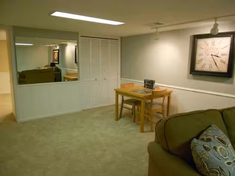 Standard living room in a senior facility with sofa, small table, clock, and mirror.