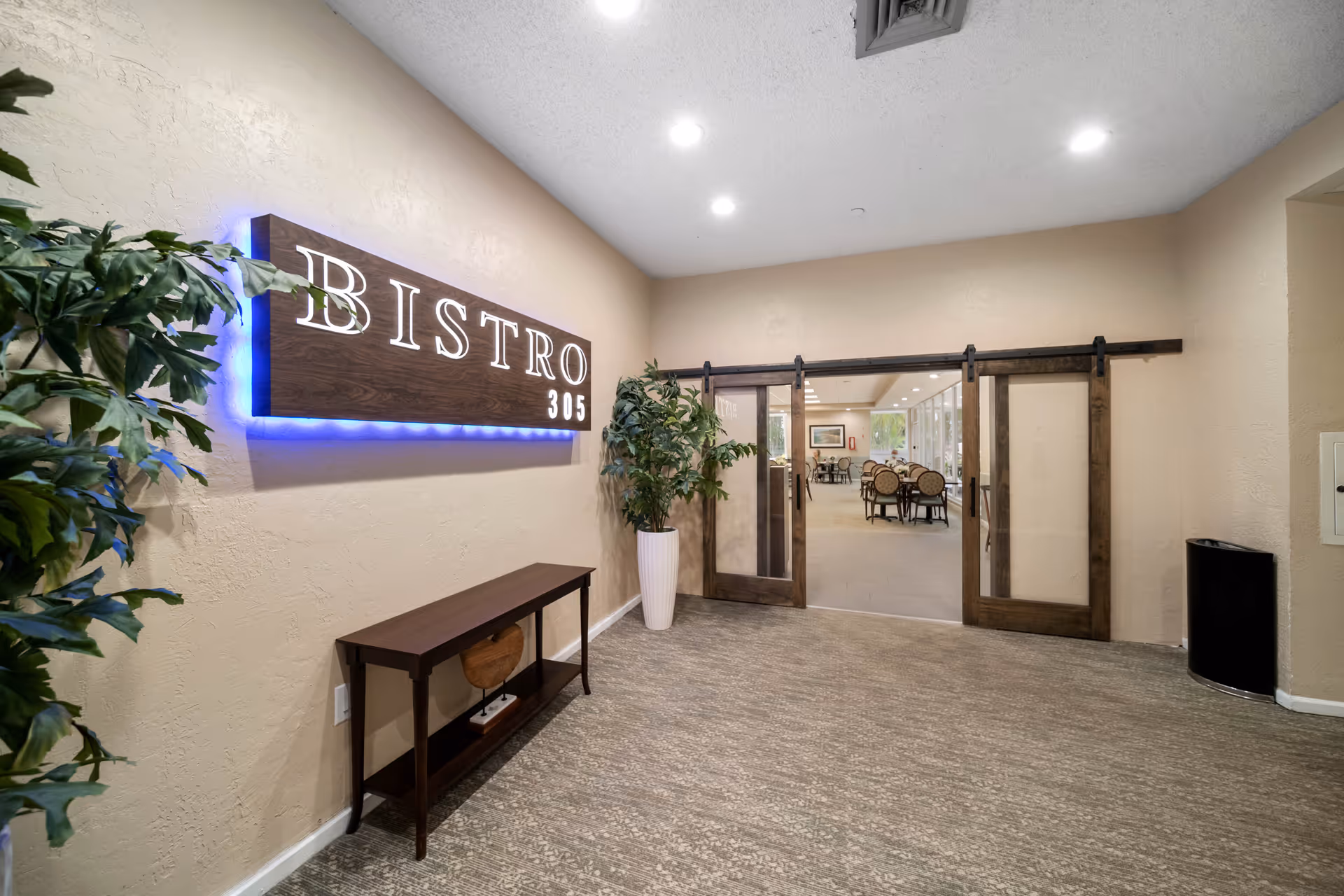 Entrance to a bistro with a wooden sign that reads 'BISTRO 305' illuminated with blue backlighting. The entrance has sliding wooden and glass doors, with a small table and potted plants on either side. Inside, tables and chairs are visible in a well-lit dining area.