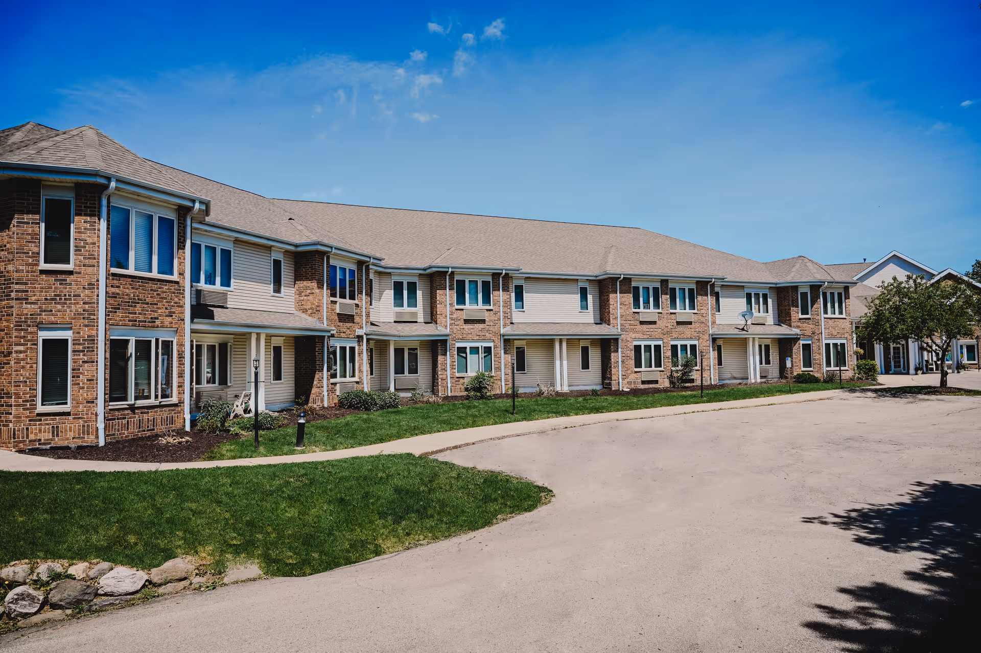 Two-story brick and beige-siding senior living building with lawns and a curved driveway under a blue sky.