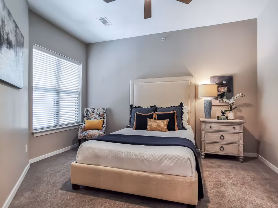 A neatly made bed with a beige upholstered headboard and a navy blue throw blanket in a bedroom. There are several pillows on the bed in shades of black, beige, and gold. To the right of the bed is a wooden nightstand with two drawers, a blue lamp, a small stack of books, and a vase with white flowers. A floral-patterned armchair with a yellow cushion is positioned near a window with white blinds. The walls are painted light gray, and the floor is carpeted in a neutral tone.