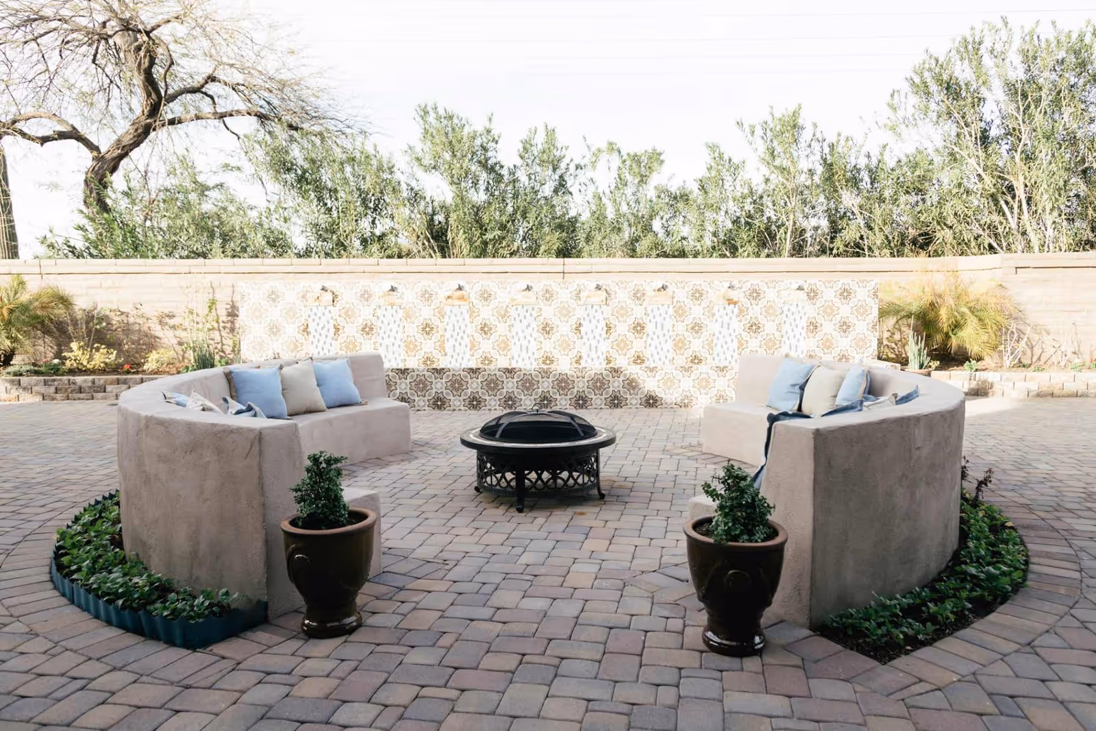 Outdoor patio area with a curved beige stucco bench adorned with blue and beige cushions surrounding a black metal fire pit. The ground is paved with stone tiles, and there are two potted plants in front of the bench. A decorative tiled wall with water spouts and greenery is visible in the background.