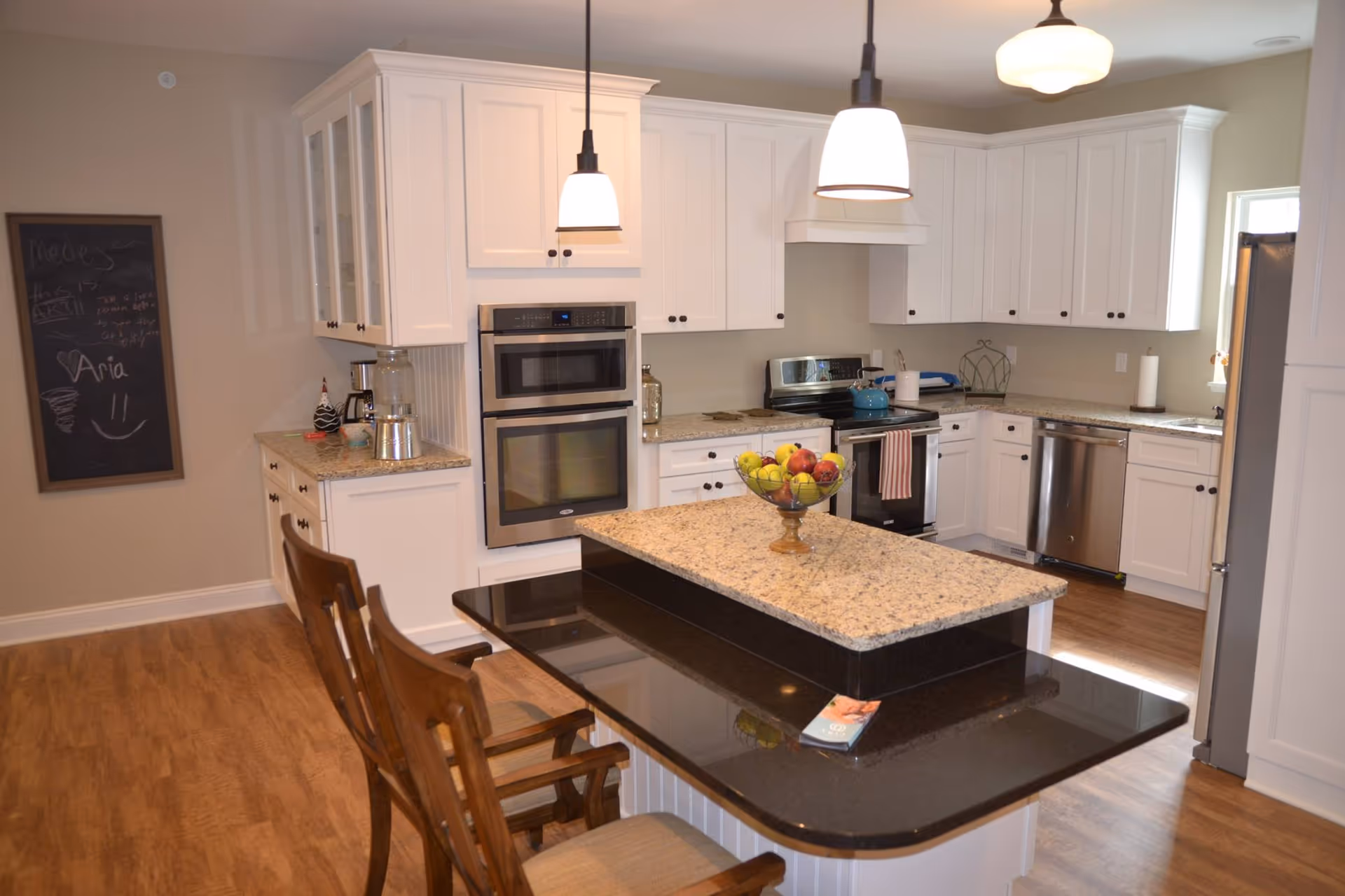 A modern kitchen with white cabinetry, granite countertops, and stainless steel appliances including a double oven, stove, dishwasher, and refrigerator. There is a kitchen island with a two-tiered countertop, two wooden chairs, and a bowl of fruit on top. Three pendant lights hang from the ceiling, and a chalkboard with writing is visible on the wall to the left.