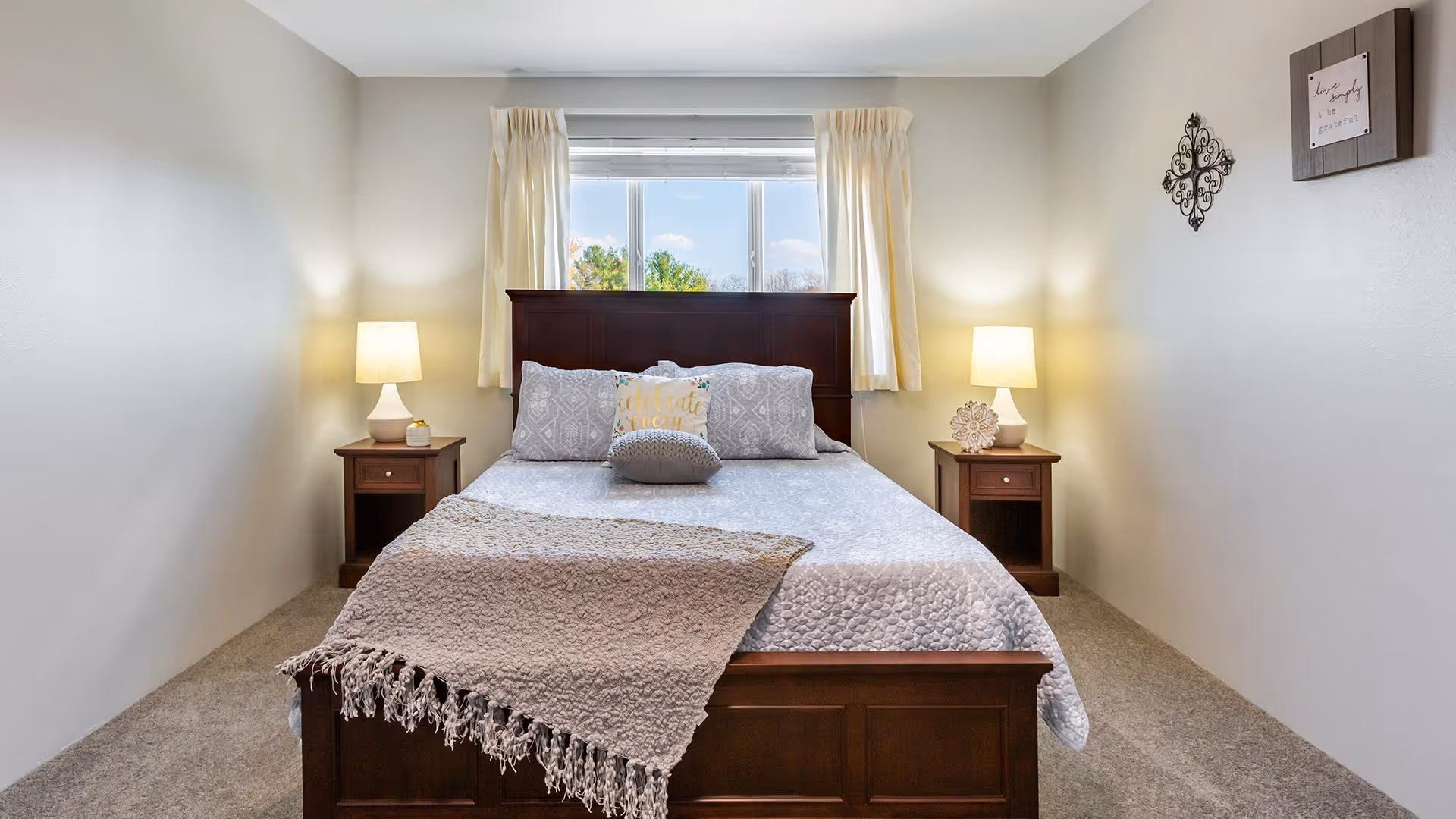 A neatly made bed with a wooden headboard and footboard is centered in a bright bedroom. The bed has a light gray quilt, decorative pillows, and a textured throw blanket draped over the foot. On each side of the bed is a wooden nightstand with a lit lamp and small decorative items. A window with cream-colored curtains is behind the bed, letting in natural light. The walls are painted light gray and have minimal wall decor.