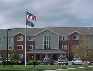 Front exterior view of a three-story senior living facility building with a combination of brick and beige siding. Two flagpoles stand in front, one with the American flag and the other with a state flag. Several cars are parked in front of the building, and there is a well-maintained lawn with small trees and shrubs.